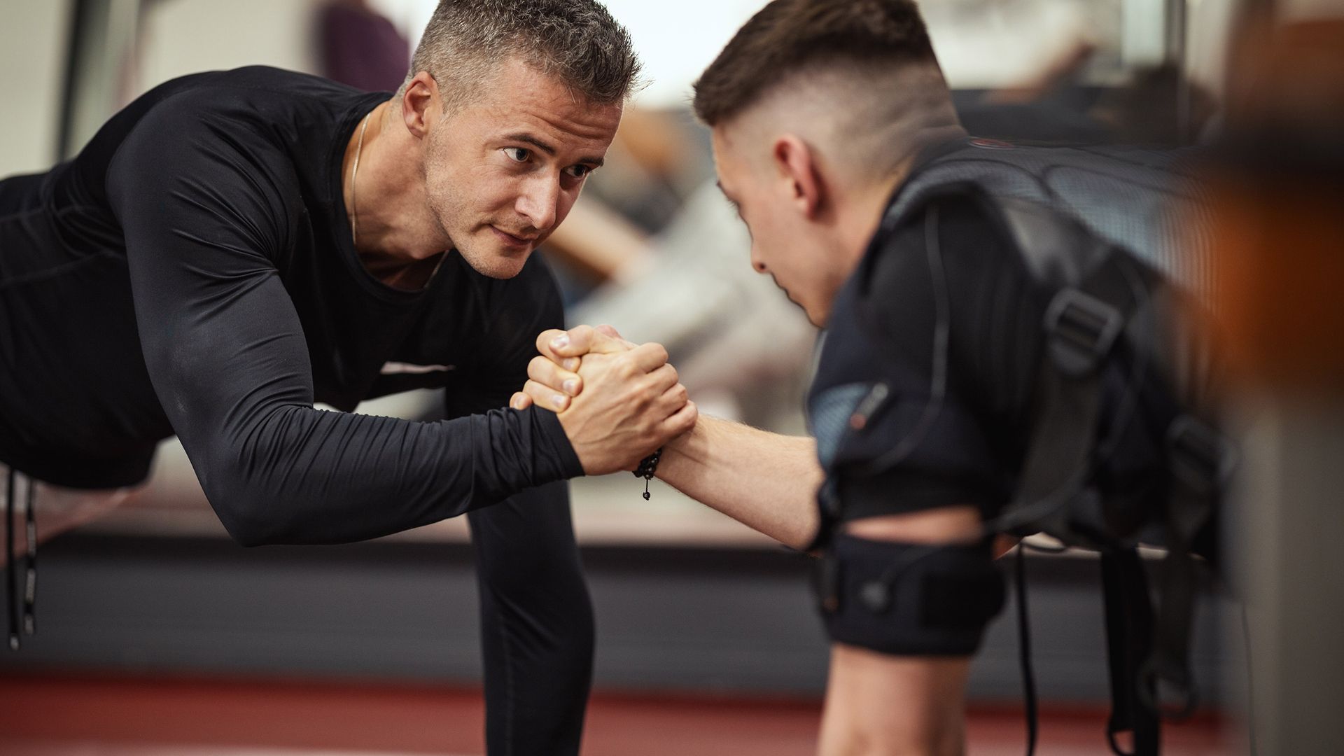 Zwei Männer in Sportkleidung messen sich im Armdrücken, einer trägt ein EMS-Trainingsgerät mit Elektroden am Oberkörper.